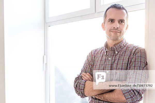 Mature man wearing check shirt leaning against window arms crossed looking away smiling