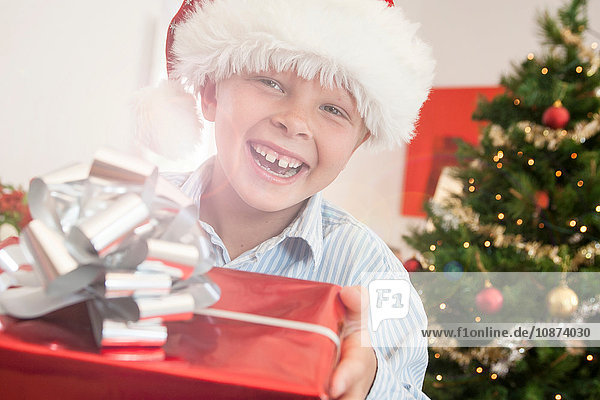 Junge mit Weihnachtsmannhut hält Weihnachtsgeschenk und schaut lächelnd in die Kamera