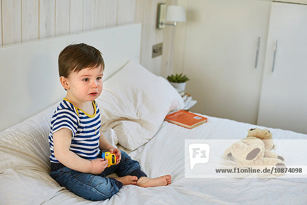 Sad baby boy sitting on bed holding toy car  looking away