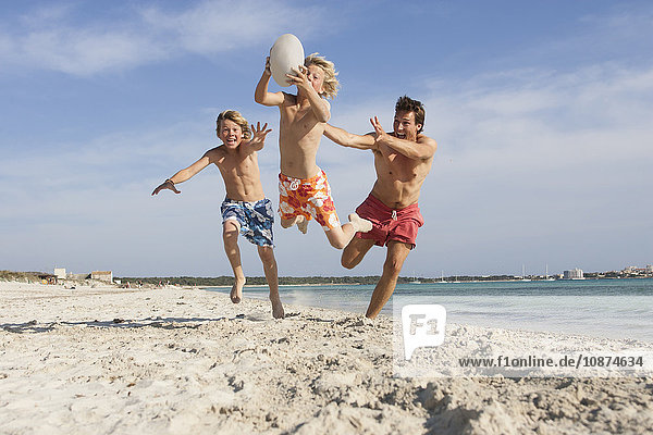 Junge springt mit Rugbyball  der von Bruder und Vater am Strand verfolgt wird  Mallorca  Spanien