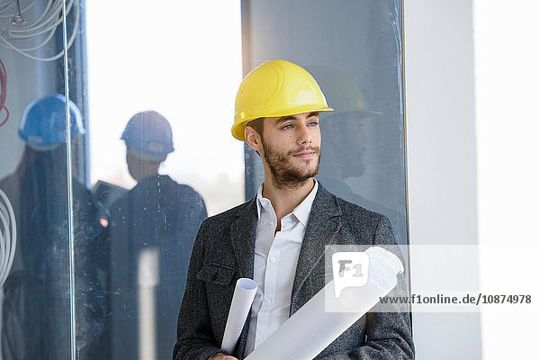Porträt eines jungen Geschäftsmannes mit gelbem Schutzhelm und Blaupause im neuen Büro