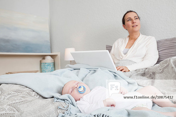 Mother sitting in bed using laptop while baby boy sleeps beside her