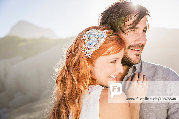Man hugging red haired woman in front of rocks looking away smiling