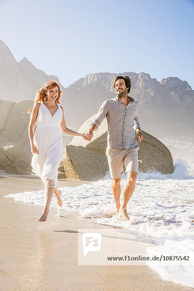 Vorderansicht in voller Länge eines am Strand laufenden Paares das sich an den Händen hält