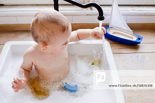 Kleiner Junge badet in Küchenspüle und berührt fließendes Wasser