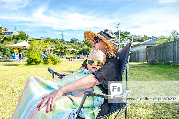 Junge mit gelber Sonnenbrille sitzt auf dem Schoß der Mutter im Park,  Waiheke Island,  Neuseeland