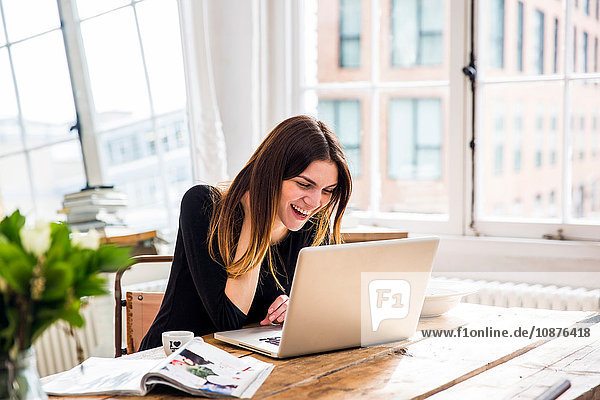 Junge Frau in Stadtwohnung lacht  während sie am Laptop arbeitet