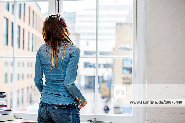 Rückansicht einer musikhörenden jungen Frau  die durch das Fenster einer Stadtwohnung schaut
