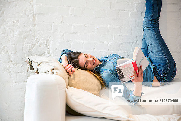 Junge Frau  die mit angehobenen Beinen auf dem Sofa liegt und ein Buch liest