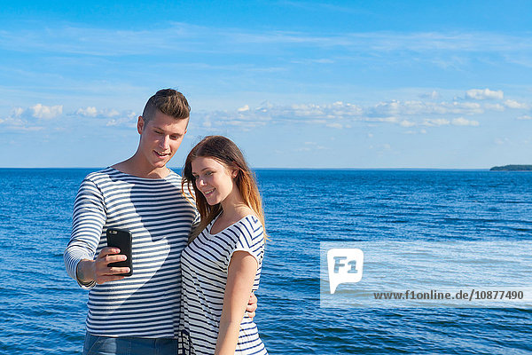 Junges Paar steht am Meer und fotografiert sich selbst mit einem Smartphone