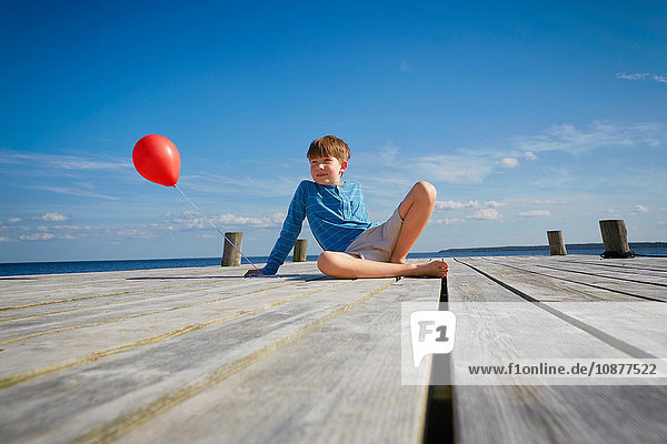 Junge sitzt auf einem Holzsteg und hält einen roten Heliumballon