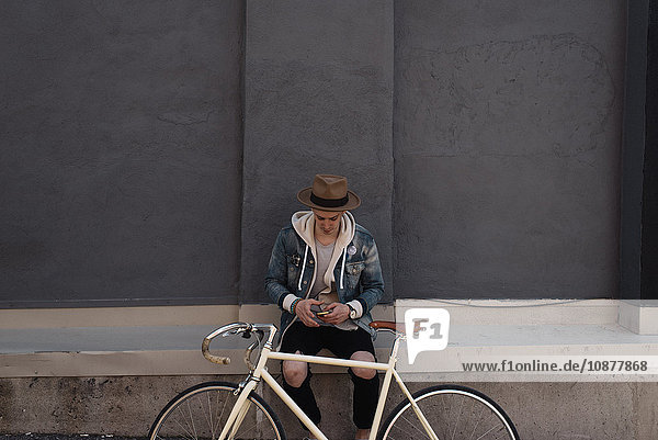 Junger Mann sitzt an der Wand  Fahrrad vor sich  benutzt Smartphone