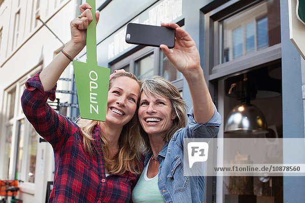 Frauen vor einem Geschäft  die ein Schild offen halten und sich mit einem Smartphone selbstständig machen