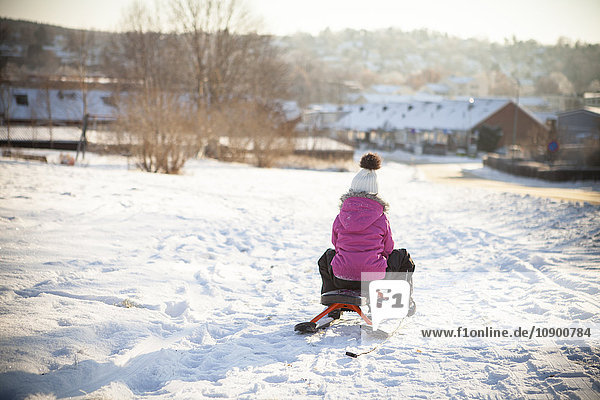 Sweden  Vastergotland  Lerum  Rear view of girl (8-9) sledding along snowy road with townscape in background
