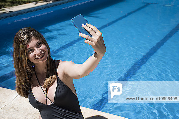 Smiling woman in swimsuit sitting at pool edge taking a selfie with smartphone