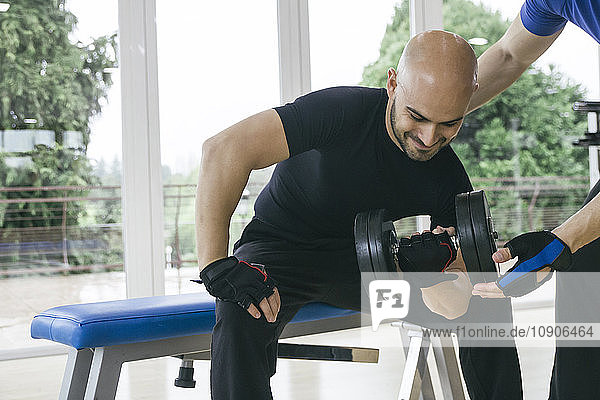 Man lifting a dumbbell sitting on a bench in a gym assisted by a trainer