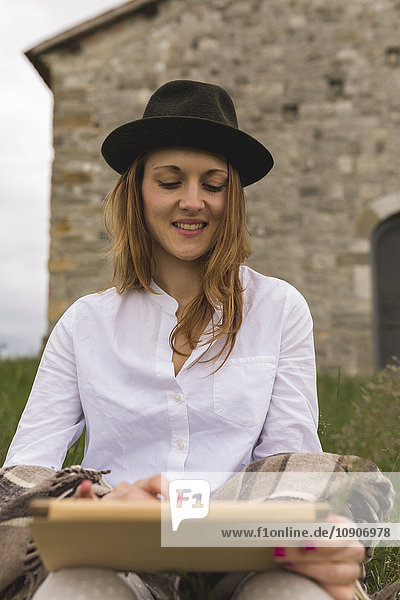 Woman with hat sitting on a meadow using digital tablet