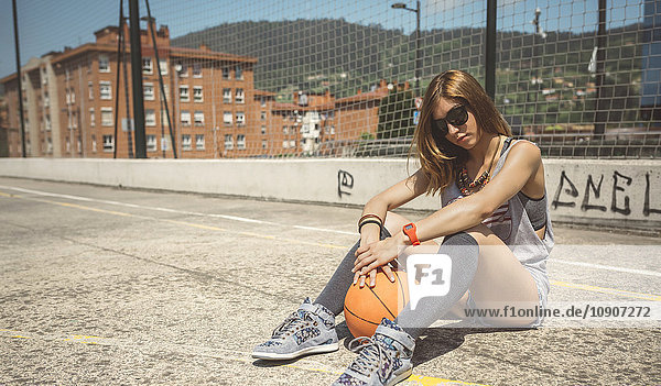 Junge Frau sitzt auf dem Boden des Baketballplatzes mit Ball zwischen den Beinen