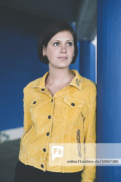 Porträt einer Frau mit gelber Jacke  die sich gegen die blaue Säule lehnt.