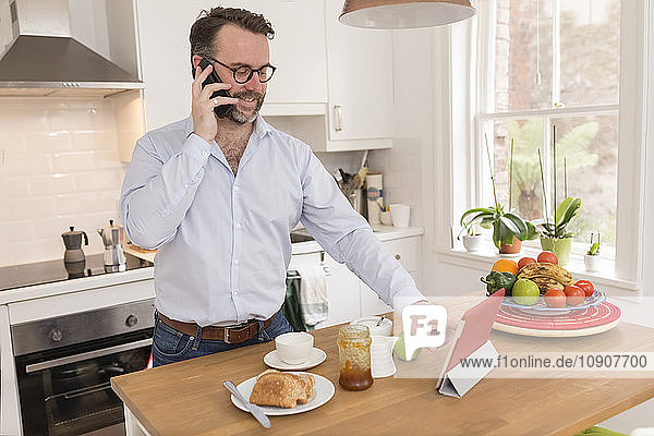 Man standing at breakfast table in the kitchen telephoning with smartphone while using digital tablet