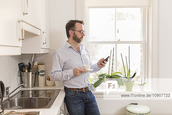 Man standing in the kitchen with cup of coffee looking at his smartphone