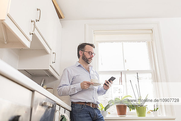 Man standing in the kitchen with cup of coffee looking at his smartphone