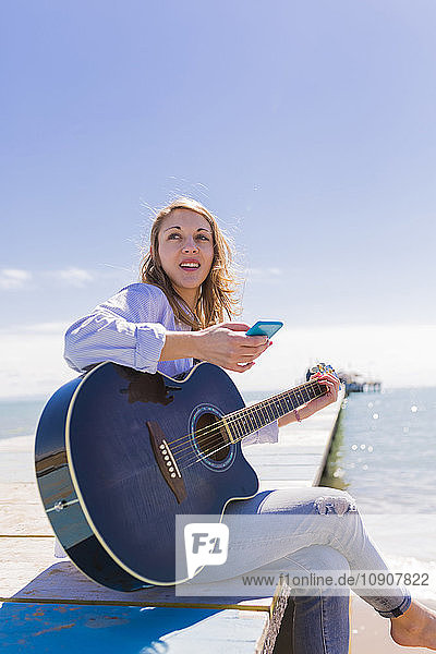 Young woman with smartphone and guitar sitting on jetty