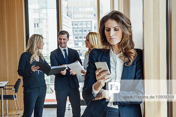 Businesswoman in office looking at cell phone with businesspeople in background