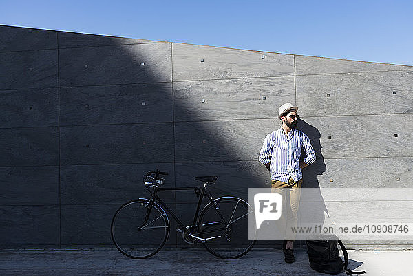Young man with bicycle and backpack leaning against wall