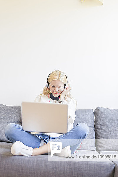 Blond young woman sitting on couch with headphones looking at laptop