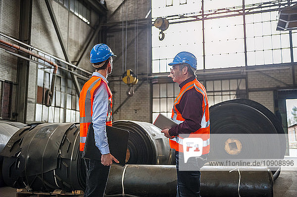 Two men with safety vests in factory hall with rolls of rubber