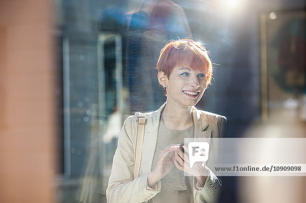 Smiling young woman holding cell phone looking at shop window