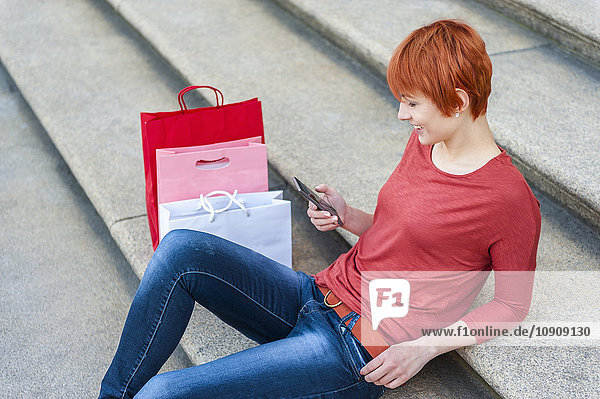 Junge Frau sitzt auf einer Treppe mit Handy und Einkaufstaschen neben ihr.