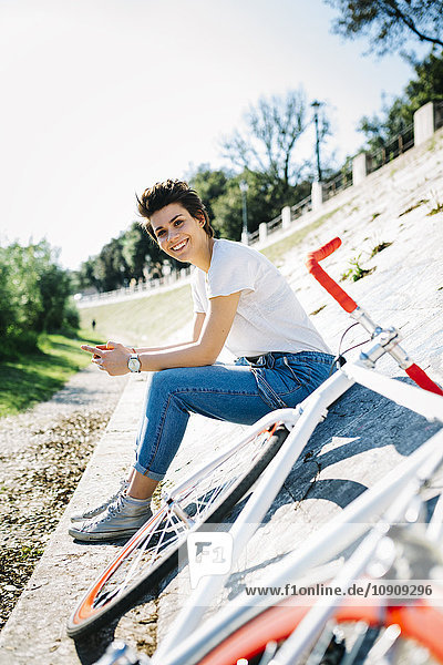 Lächelnde junge Frau mit Fahrrad auf geneigter Wand sitzend