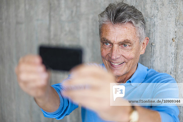 Lächelnder älterer Mann an der Betonwand  der einen Selfie nimmt.