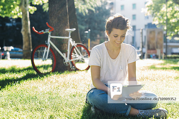 Lächelnde junge Frau auf der Wiese sitzend mit digitalem Tablett