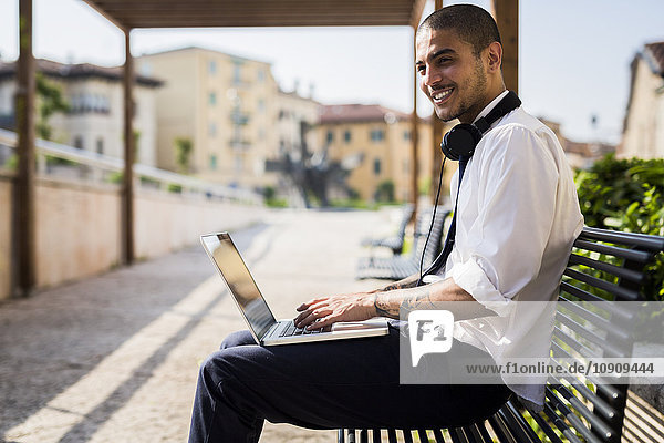 Smiling young businessman sitting on a bench using laptop