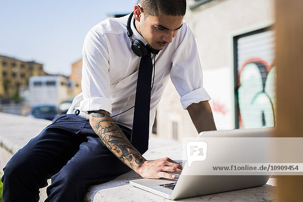 Junger Geschäftsmann sitzt auf einer Wand mit Laptop