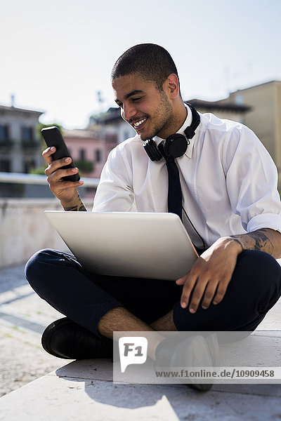 Smiling young businessman sitting on a wall with laptop looking at his cell phone