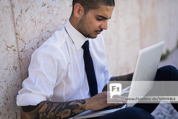 Serious young businessman sitting on the ground using his laptop