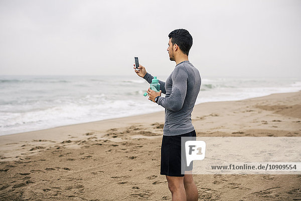 Sportive young man with cell phone and drinking bottle on the beach