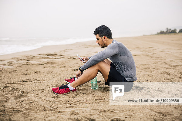 Sportlicher junger Mann mit Handy und Trinkflasche am Strand