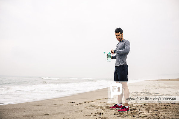 Sportlicher junger Mann mit Trinkflasche am Strand mit Blick auf die Uhr