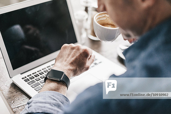 Reifer Mann  der Kaffee trinkt  während er am Laptop arbeitet  mit Blick auf smartwatch