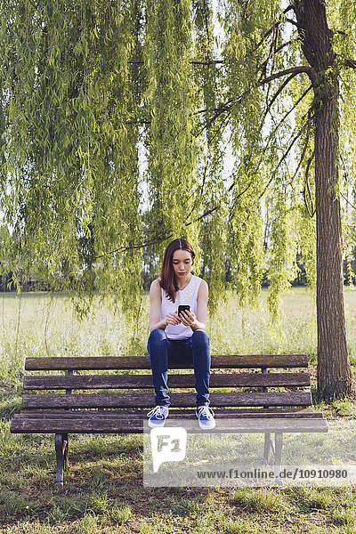 Junge Frau mit Telefon sitzt auf einer Bank im Park