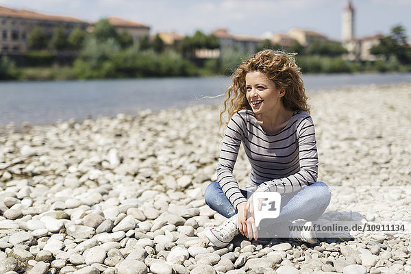 Italy  Verona  portrait of happy woman sitting on stony beach at riverside