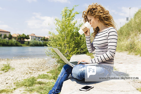 Frau trinkt Kaffee  um zu gehen  während sie den Laptop am Flussufer benutzt.