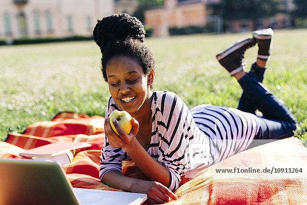 Frau  die auf einer Wiese auf einer Decke liegt und Apfel isst.
