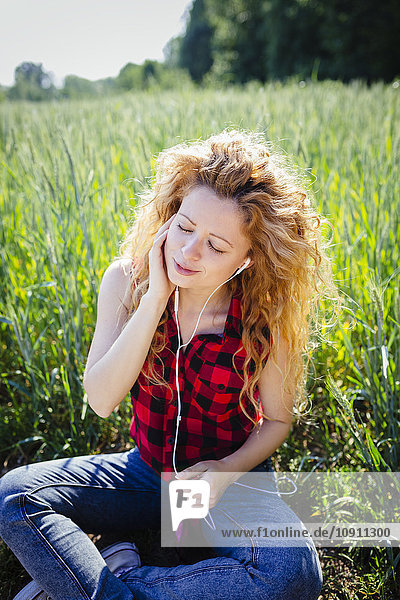 Porträt einer Frau  die in einem Feld sitzt und Musik mit Kopfhörern hört.