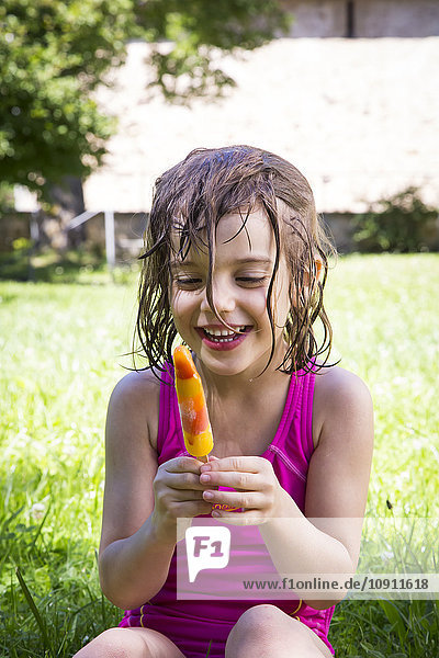 Porträt eines glücklichen kleinen Mädchens mit nassen Haaren  das auf einer Wiese mit Eis am Stiel sitzt.